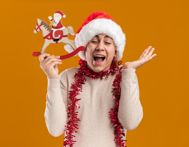 Animado com os olhos fechados, jovem usando chapéu de Natal com guirlanda no pescoço, segurando um brinquedo de Natal, espalhando a mão isolada na parede amarela