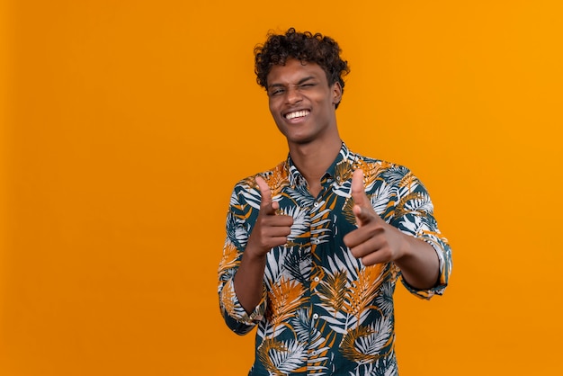 Animado e positivo jovem bonito de pele escura com cabelo encaracolado em uma camisa estampada de folhas olhando feliz enquanto aponta para a câmera com dois dedos