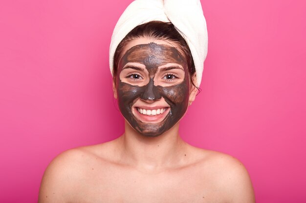 Animado feliz fêmea posa com sorriso e chocolate máscara facial, com ombros nus, cuida de sua beleza e aparência, usa toalha branca na cabeça, isolada sobre parede rosa.