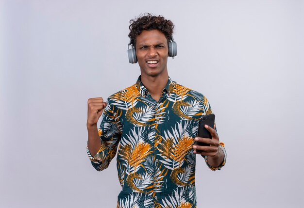 Animado, jovem, bonito, de pele escura, com cabelo encaracolado, camisa estampada de folhas em fones de ouvido, segurando um smartphone com o punho cerrado