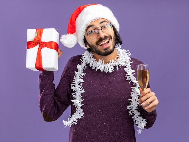 Animado jovem bonito usando chapéu de Natal com guirlanda no pescoço e segurando uma caixa de presente com taça de champanhe isolada na parede azul