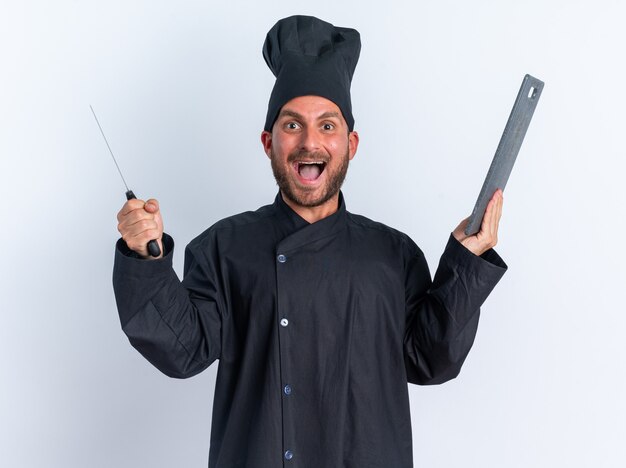 Animado jovem cozinheiro masculino caucasiano com uniforme de chef e boné segurando o cutelo e a tábua de cortar, olhando para a câmera isolada na parede branca