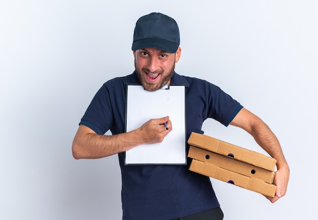 Foto grátis animado jovem entregador caucasiano de uniforme azul e boné segurando pacotes de pizza e prancheta sob o queixo olhando para a câmera apontando para a prancheta com uma caneta isolada na parede branca