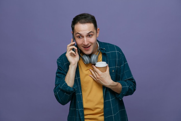 Animado jovem estudante do sexo masculino usando fones de ouvido no pescoço segurando uma xícara de café de papel apontando para si mesmo com a mão olhando para baixo enquanto fala no telefone isolado no fundo roxo