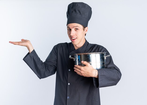 Animado jovem loiro cozinheiro masculino com uniforme de chef e boné segurando a panela, mostrando a mão vazia