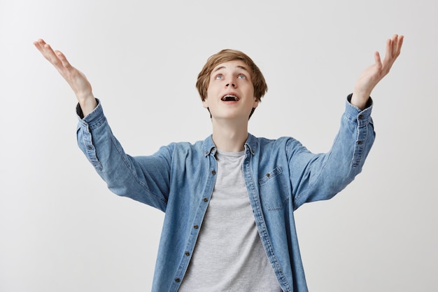 Animado jovem loiro em roupas jeans feliz que amigos vieram na festa, levanta as mãos com prazer, feliz em receber parabéns e presentes, comemora evento especial em sua vida