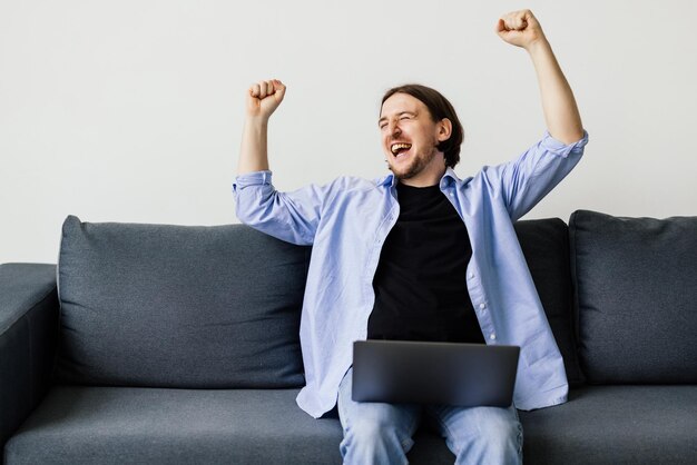 Animado jovem vencedor usando computador portátil enquanto está sentado em um sofá em casa