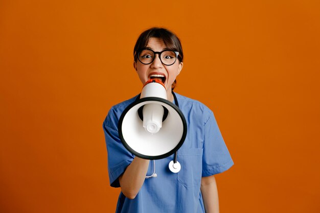 Animado segurando alto-falante jovem médica usando estetoscópio uniforme de quinto isolado em fundo laranja