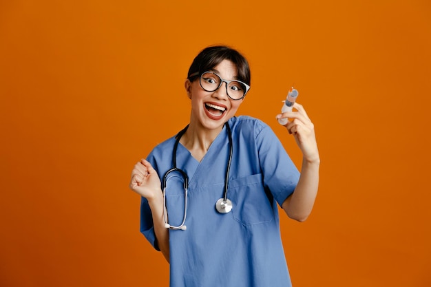 Animado segurando seringa jovem médica usando estetoscópio uniforme de quinto isolado em fundo laranja