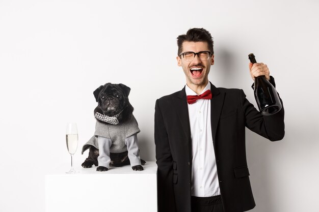 Animais de estimação, férias de inverno e conceito de ano novo. Homem feliz comemorando a festa de Natal do animal de estimação, em pé com o cachorro fofo fantasiado, bebendo champanhe e regozijando-se, fundo branco