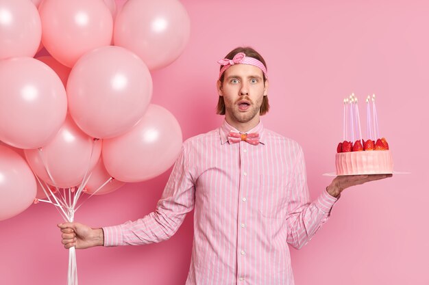 Aniversariante surpreso e chocado ao receber tantos parabéns de amigos e parentes posa com balões e bolo festivo vestido com tiara de camisa borboleta isolada sobre parede rosa