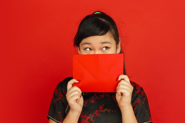 Ano novo chinês 2020. retrato de jovem asiático isolado sobre fundo vermelho. modelo feminino com roupas tradicionais parece um sonho e apresentando envelope vermelho. celebração, feriado, emoções.