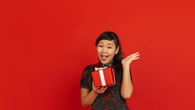 Ano Novo Chinês. Retrato de jovem asiático isolado sobre fundo vermelho. Modelo feminino com roupas tradicionais parece feliz, sorridente e surpreso com a caixa de presente. Celebração, feriado, emoções.