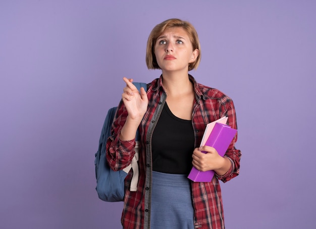 Ansiosa aluna eslava usando uma mochila cruzando os dedos segurando um livro e um caderno