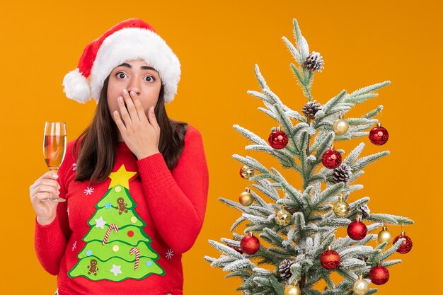 ansiosa jovem caucasiana com chapéu de Papai Noel coloca a mão na boca e segura a taça de champanhe ao lado da árvore de Natal isolada em um fundo laranja com espaço de cópia