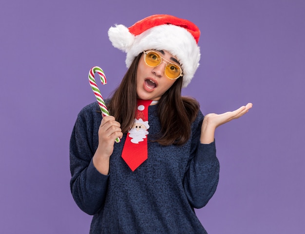 Ansiosa jovem caucasiana de óculos de sol com chapéu de papai noel e gravata de papai noel segurando o bastão de doces e mantém a mão aberta isolada no fundo roxo com espaço de cópia