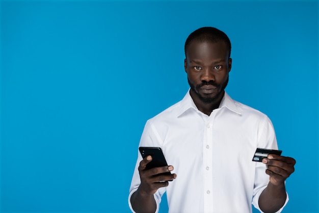 Ansioso homem afro-americano está segurando o cartão de crédito e o celular nas mãos