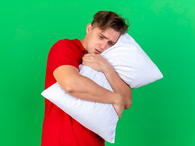 Ansioso jovem bonito loiro doente abraçando o travesseiro, olhando para baixo, isolado na parede verde