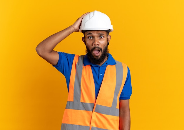 Ansioso jovem construtor afro-americano de uniforme com capacete de segurança, colocando a mão na cabeça, isolado em parede laranja com espaço de cópia