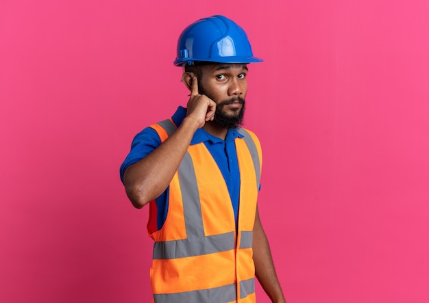 ansioso jovem construtor de uniforme com capacete de segurança colocando o dedo na orelha isolado na parede rosa com espaço de cópia