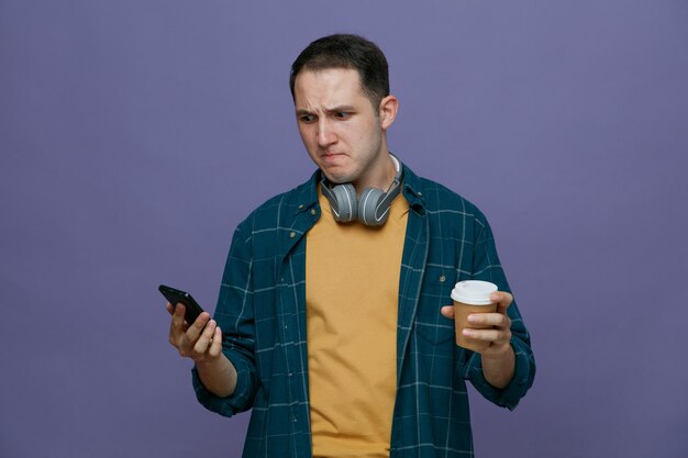 Ansioso jovem estudante do sexo masculino usando fones de ouvido no pescoço, segurando a xícara de café de papel e o celular olhando para o celular isolado no fundo roxo