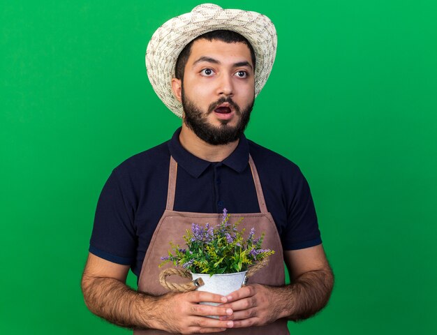 ansioso jovem jardineiro caucasiano com chapéu de jardinagem, segurando o vaso de flores e olhando para o lado isolado na parede verde com espaço de cópia