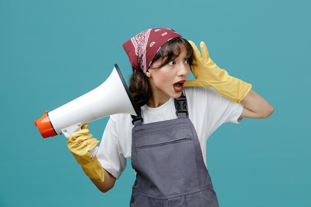 Ansioso jovem limpador feminino vestindo bandana uniforme e luvas de borracha segurando alto-falante olhando para o lado, mantendo a mão na cabeça isolada em fundo azul