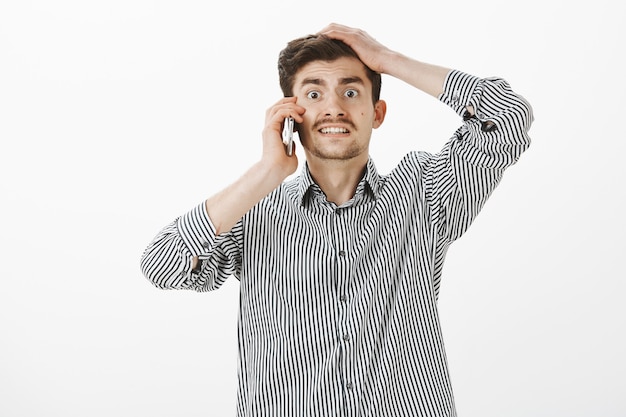 ansioso, preocupado, engraçado, europeu, com barba e bigode, fazendo cara de nervosismo culpado e segurando a mão na cabeça enquanto falava no smartphone, chegando atrasado e inventando uma desculpa