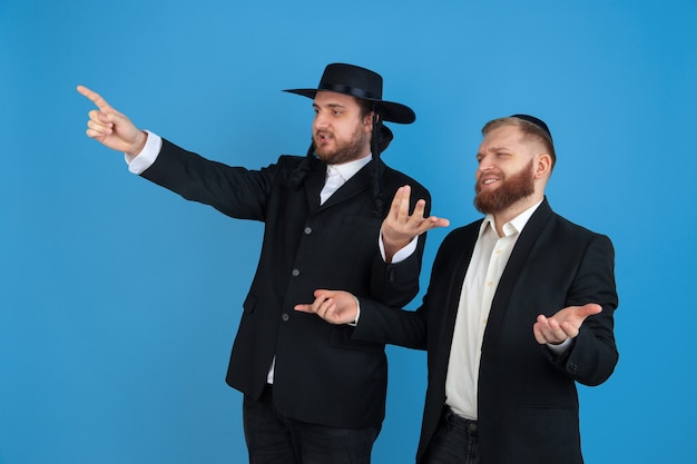 Foto grátis apontando, convidativo. retrato de um jovem judeu ortodoxo isolado na parede azul. purim, negócios, festival, feriado, celebração pessach ou páscoa, judaísmo, conceito de religião.