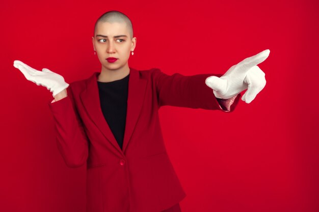 Apontando, mostrando. Retrato de uma jovem mulher careca caucasiana, isolada na parede vermelha.