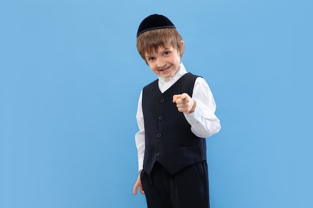 Apontando. Retrato de um jovem rapaz judeu ortodoxo isolado na parede azul. Purim, negócios, festival, feriado, infância, celebração Pessach ou Páscoa, judaísmo, conceito de religião.