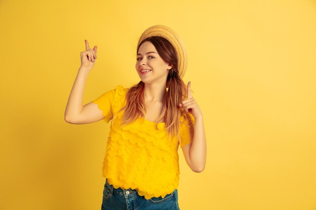 Apontando, sorrindo. Retrato de uma mulher caucasiana em fundo amarelo do estúdio. Bela modelo feminino no chapéu. Conceito de emoções humanas, expressão facial, vendas, anúncio. Verão, viagens, resort.