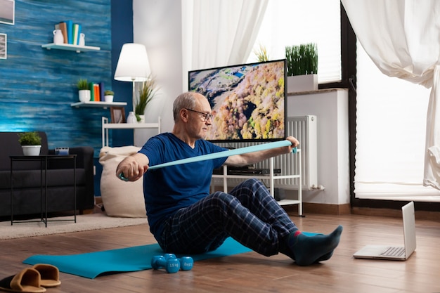 Aposentado trabalhando na resistência do corpo, exercitando os músculos dos braços usando uma faixa elástica, sentado no tapete de ioga com as pernas cruzadas. Homem sênior fazendo exercícios durante a aula de ginástica, olhando no laptop