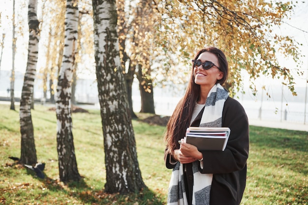 Apreciando a natureza e dias quentes. jovem morena sorridente em óculos de sol fica no parque perto das árvores e mantém o bloco de notas