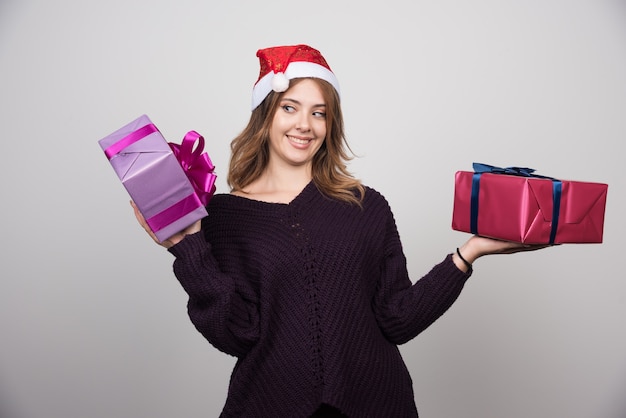 Apresenta-se jovem mulher com chapéu de Papai Noel segurando caixas de presente.