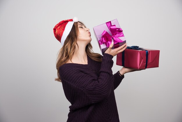 Apresenta-se jovem mulher com chapéu de Papai Noel segurando caixas de presente.