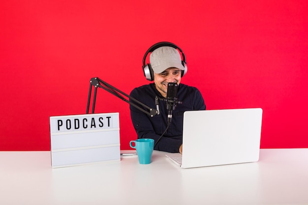 Apresentador masculino com boné em estúdio de gravação de rádio podcast sorrindo e consultando a internet, ao lado de um computador, um microfone e uma caixa de luz com a palavra podcast, sobre fundo vermelho.