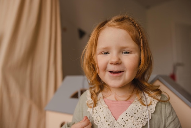 Aproximação de uma linda garotinha caucasiana com cabelo vermelho olha para a câmera no quarto Conceito de crianças