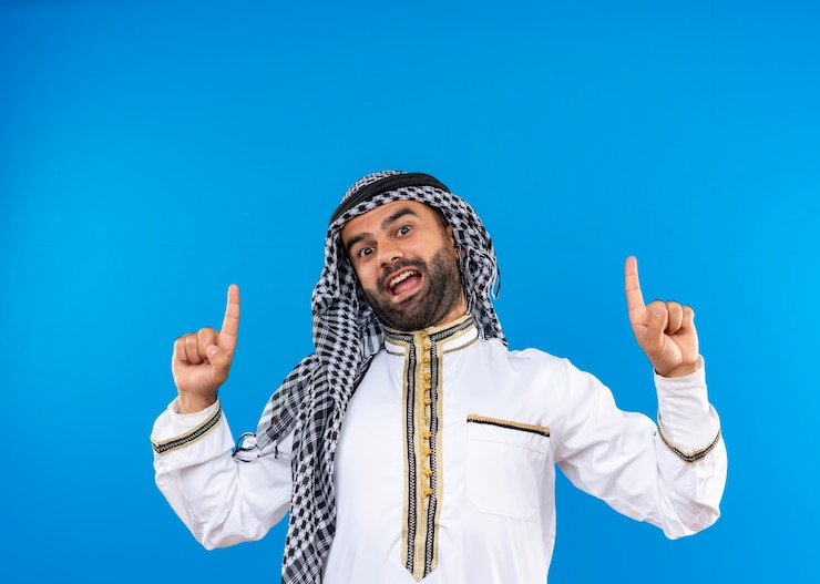 Foto grátis árabe com roupa tradicional apontando com os dedos para cima e sorrindo alegremente em pé sobre a parede azul