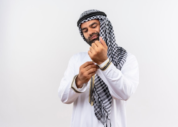 Árabe, com roupa tradicional, consertando a abotoadura com ar confiante em pé sobre uma parede branca