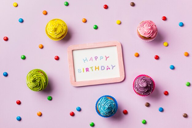 Ardósia de feliz aniversário rodeada de gemas coloridas e muffins em fundo rosa