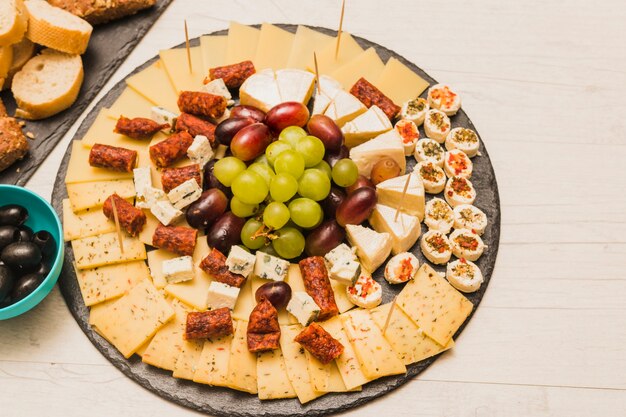 Ardósia preta circular com prato de queijo; uvas e enchidos na mesa de madeira
