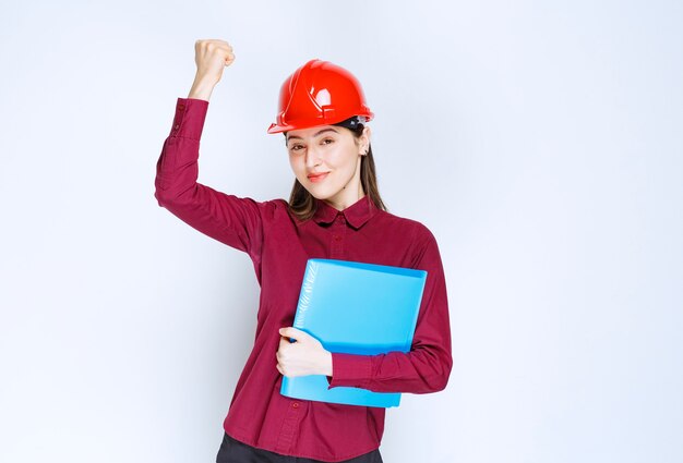 Arquiteta feminina no capacete vermelho, segurando uma pasta azul com documentos.