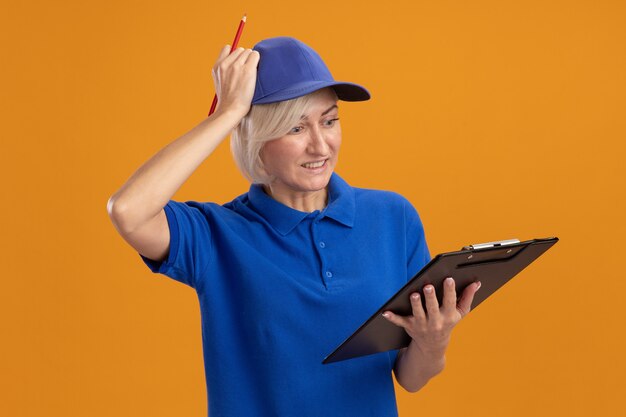 arrependendo-se da entregadora loira de meia-idade com uniforme azul e boné segurando uma prancheta e um lápis olhando para a prancheta colocando a mão na cabeça isolada em um fundo laranja