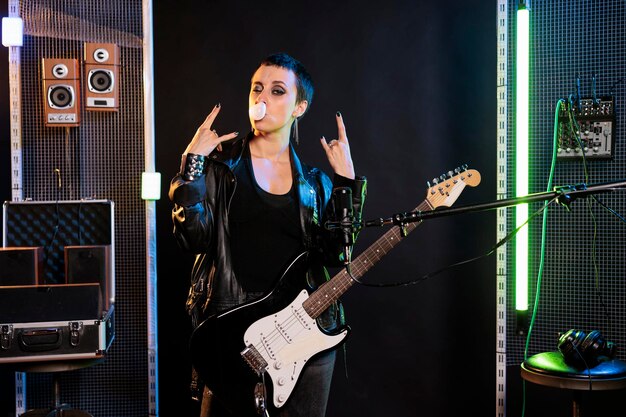 Foto grátis artista de mulher soprando chiclete enquanto posava no estúdio fazendo o gesto de estrela do rock, preparando a guitarra elétrica antes de realizar o concerto de metal. músico rebelde tocando para performance solo