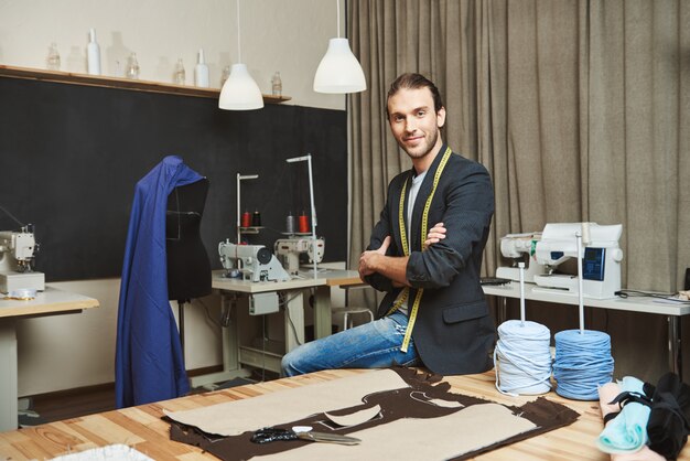 Artista e seu local de trabalho. Retrato do designer de roupas masculino caucasiano atraente maduro com penteado elegante e roupa elegante, sentado em seu estúdio, posando para a foto na revista de moda.