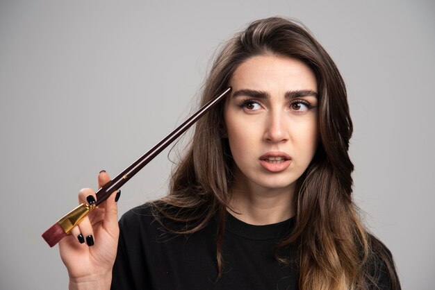 Artista feminina em camisa preta, tocando sua cabeça com o pincel.