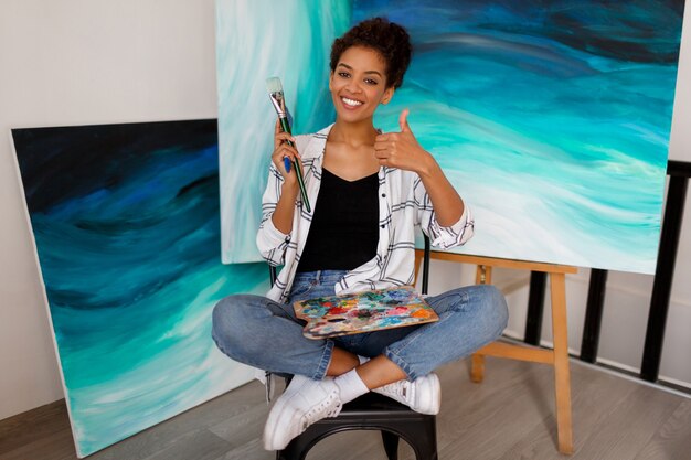 Artista feminina engraçada sentado com incrível arte abstrata de acrílico mar desenhado à mão. Segurando pincéis e paleta