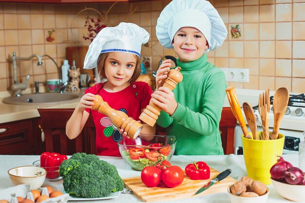 As crianças engraçadas da família feliz estão preparando uma salada de legumes fresca na cozinha