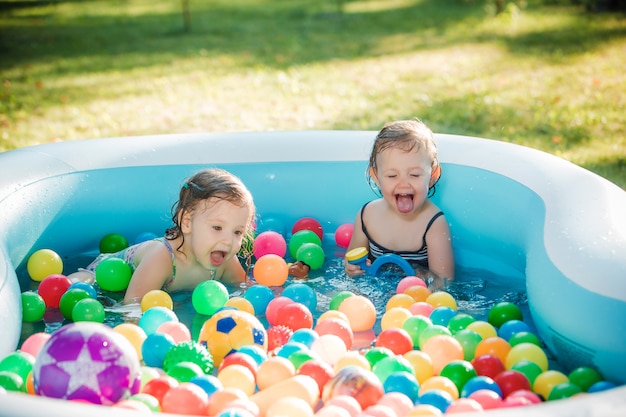 As duas meninas de dois anos de idade brincando com brinquedos na piscina inflável no dia ensolarado de verão